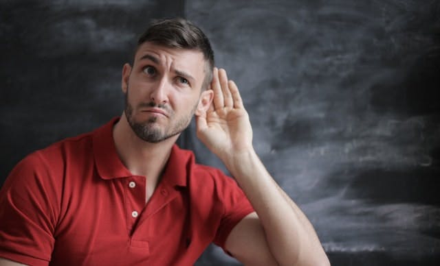 man in red shirt against black background with his hand up to his ear as if listening