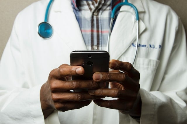 close up of a doctor from the shoulders to the waist. they have a stethoscope hanging around their neck and they're using a mobile phone