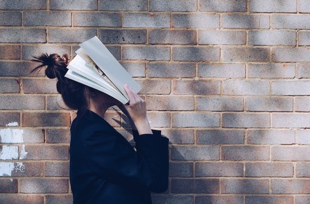 person with open book held against their face, standing by a brick wall