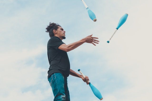 shot from low angle of someone juggling 3 blue juggling batons. behind them is a blue sky with clouds