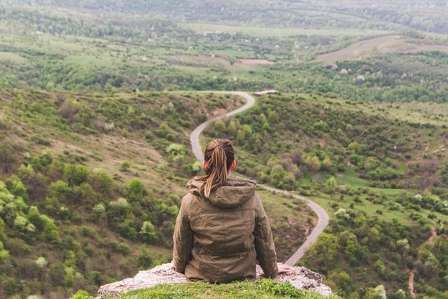 person sitting at edge of a rock, back to camera, overlooking a road stretching into the distance