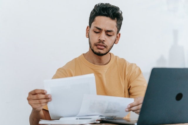 person sitting in front of laptop looking down at pieces of paper in their hands
