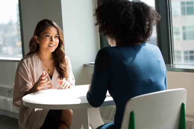 two people seated and talking at a round, sunlit table in an office setting