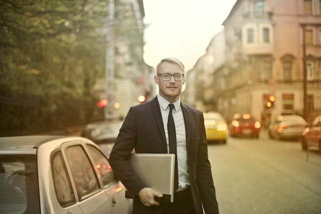 person stands in business attire is standing in the street outside of a car, looking into the camera. They have documents under their arm.