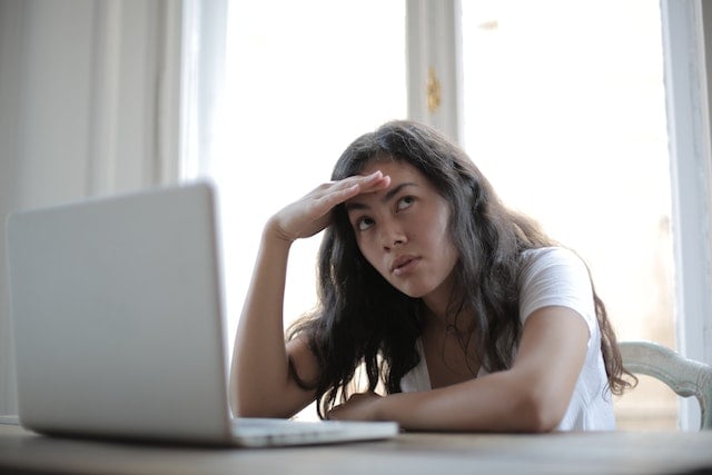 person seated at laptop in a brightly lit room, looking upwards with their hand on their head