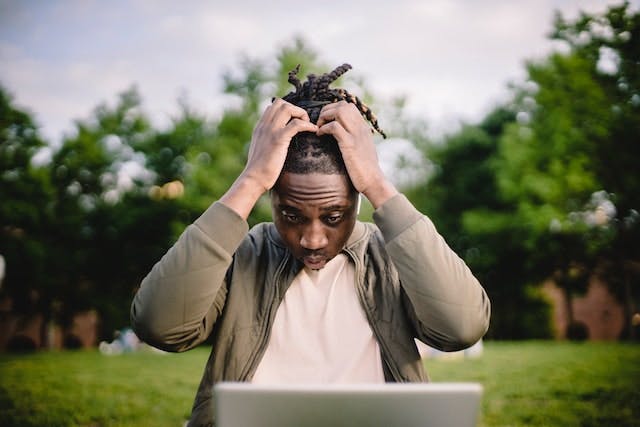 person seated outside in a park with a laptop. the person is in focus, looking down at the laptop screen and holding their head with a worried look on their face