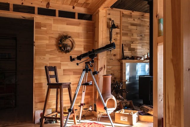 A black telescope stands in a cozy room with wooden walls, floors, and chair