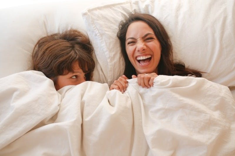 two women next to each other in bed. one is laughing, the other is peeking out from behind the covers at her
