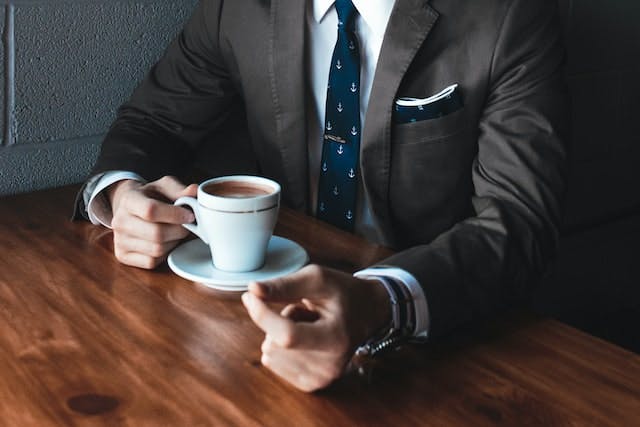 close shot of a person's torso. this person is sitting at a table, wearing a suit, with their hand on a mug of coffee. they are making a sign with their hand to indicate money