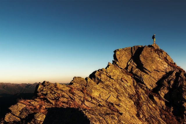 Person on the peak of a mountain overlooking the view. The sky is a deep blue behind them, fading into orange