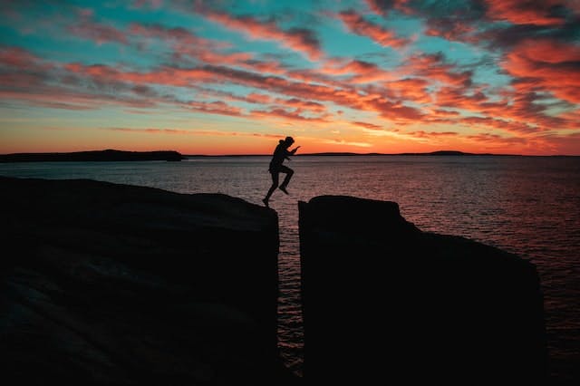 silhouette of someone jumping over a gap from one cliff to another, with the sea and sunset in the background
