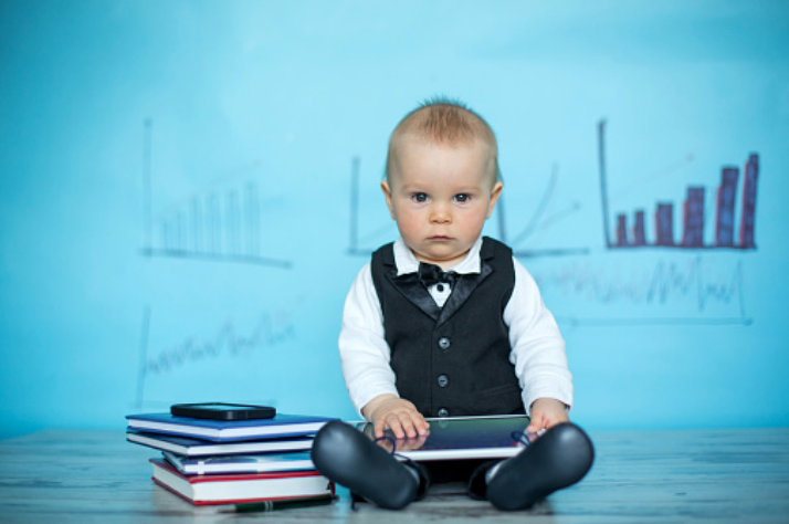 baby in a suit shirt and vest sits next to a pile of books, with a smartpad on their lap, and graphs drawn on a whiteboard in the background