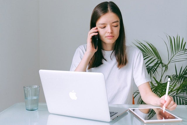 person on phone in a room with white walls, a white table, and a plant in the background. they are seated at a table, phone in one hand, taking notes on a tablet with the other, with a laptop and glass of water in front of them