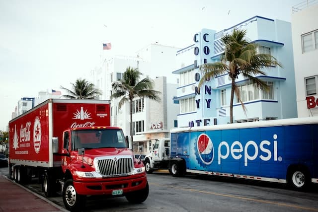 a Coca-cola semi-truck next to a Pepsi semi-truck on a road