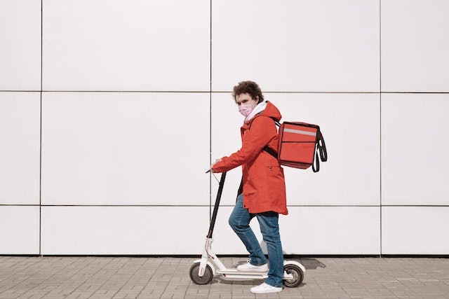 person on an electric scooter against a white wall. They are wearing a red coat and a red food delivery backpack
