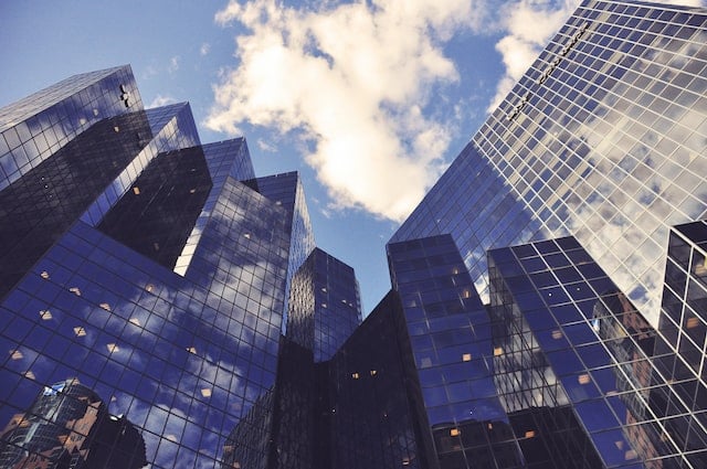 picture of mirrored skyscrapers from the ground, and angled-up photo. there is a blue sky with white clouds above the buildings